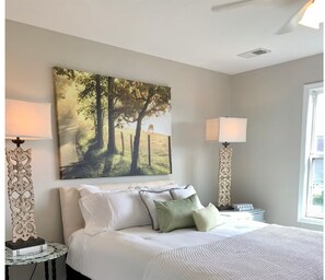 Ein Schlafzimmer mit einem Bett, zwei Nachttischlampen, einem Wandbild und einem Fenster.