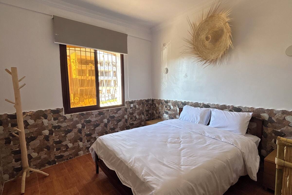 Un dormitorio con cama, pared de piedra, ventana con barrotes y l ámpara en el techo.