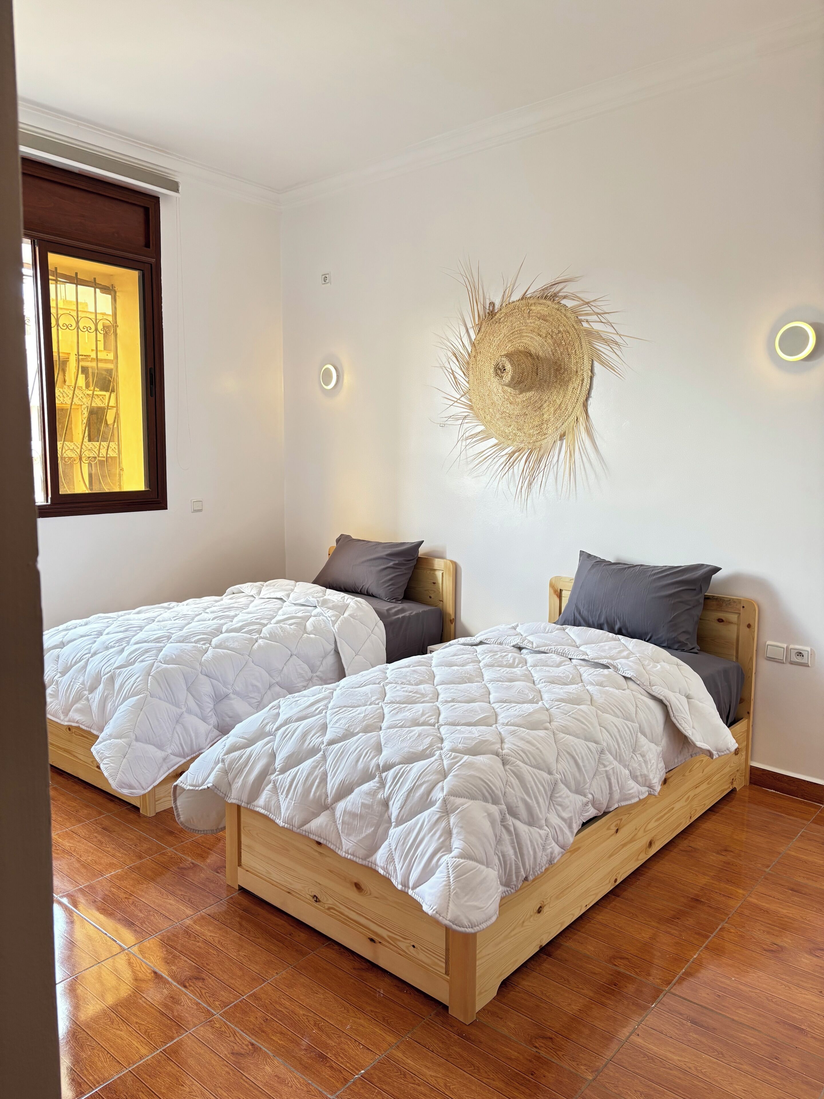 Un dormitorio con dos camas de madera, ropa de cama blanca, un adorno de pared de paja y una ventana con vistas.