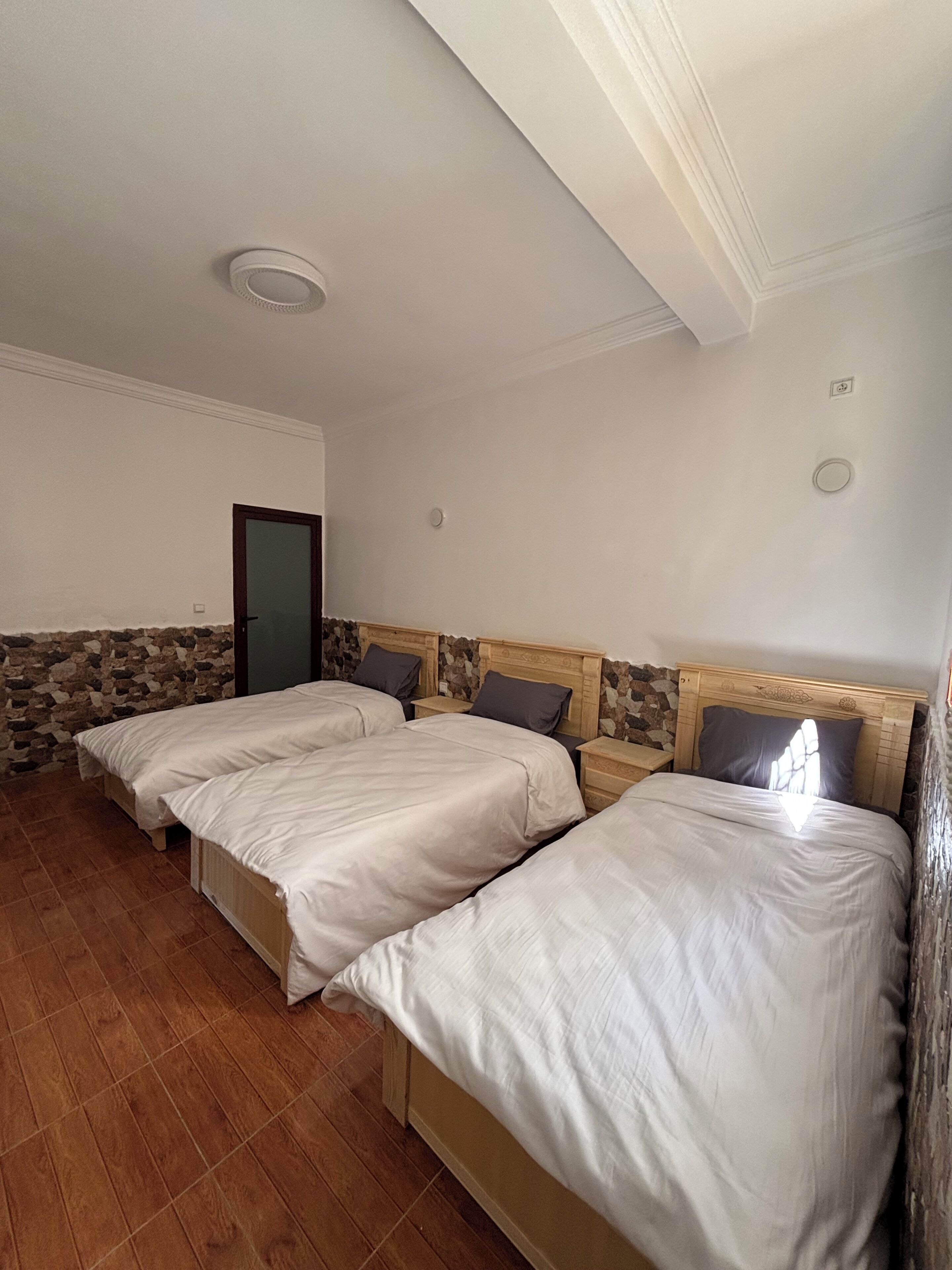 Una habitación con dos camas, una pared de piedra, cabecero de madera, una puerta y una lámpara en el techo.