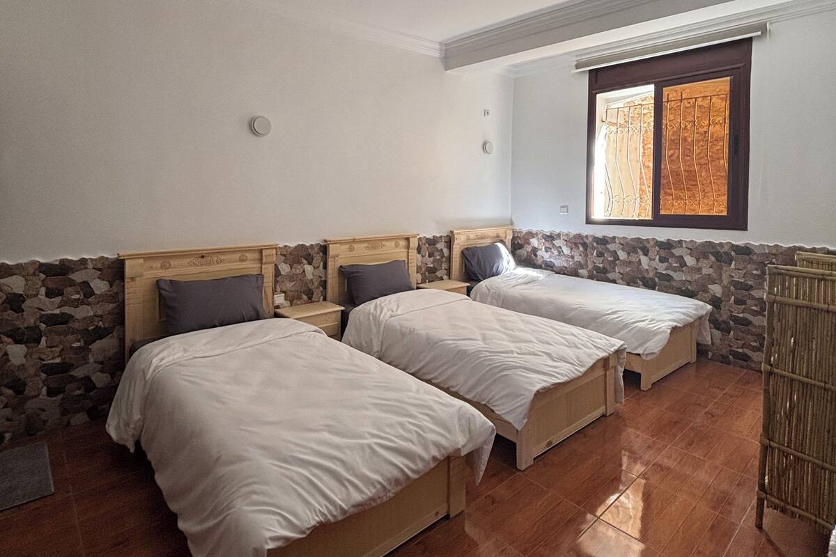 Habitación con dos camas, pared de piedra, ventana con vistas y una lámpara en el techo.