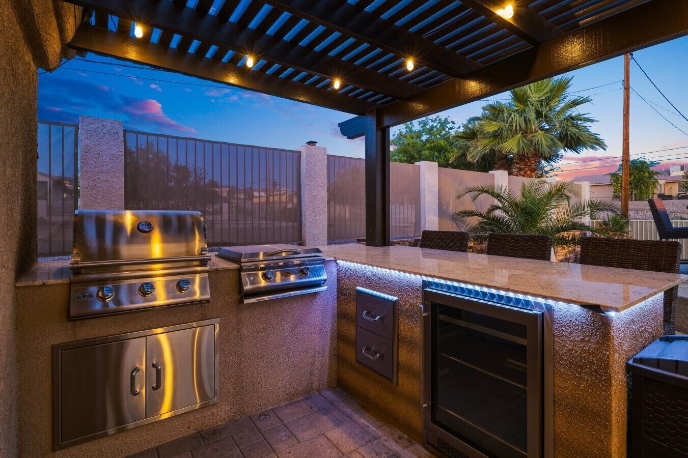 An outdoor kitchen with a grill, oven, and bar area, under a covered patio with a view of the sunset.