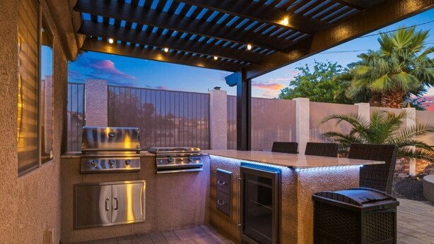 Une cuisine extérieure équipée d’un barbecue, d’un four et d’un réfrigérateur encastré, située sous une pergola illuminée de guirlandes lumineuses.