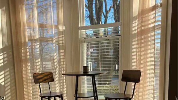 Una habitación con dos sillas y una mesa, una ventana con persianas y vistas a árboles y edificios.