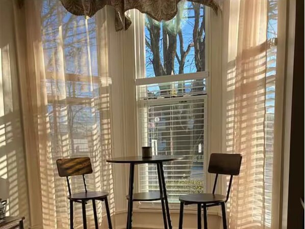 Ein Zimmer mit zwei Stühlen, einem Tisch, einem Fenster mit Jalousien und Blick auf Bäume und Gebäude.