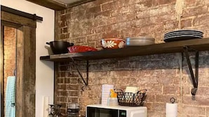 Una cocina con pared de ladrillo, microondas, repisa con vajilla y una cesta.