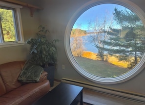 A room with a brown leather sofa, a potted plant, a round window with a view of trees and a body of water.