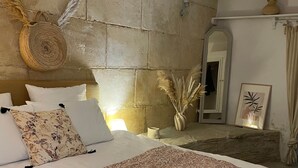 Quarto com parede de pedra, cama com roupa de cama estampada, cesta de palha e um vaso com plumas de capim-do-avelar.