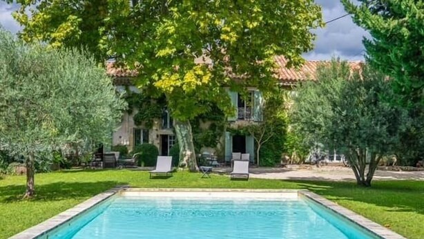 Piscina com espreguiçadeiras, rodeada por árvores e um edifício com telhado de telhas vermelhas.