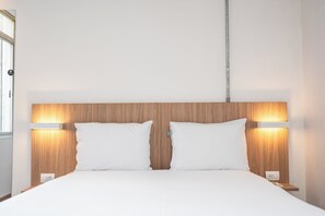 Habitación de hotel con cabecera de madera, dos almohadas blancas, lámparas de noche y mesita con toma de corriente.