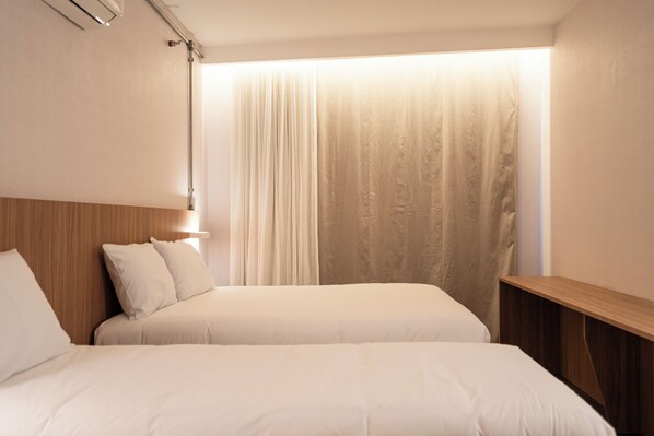 A hotel room with two beds, a wooden headboard, a bedside table, and a window with sheer curtains.