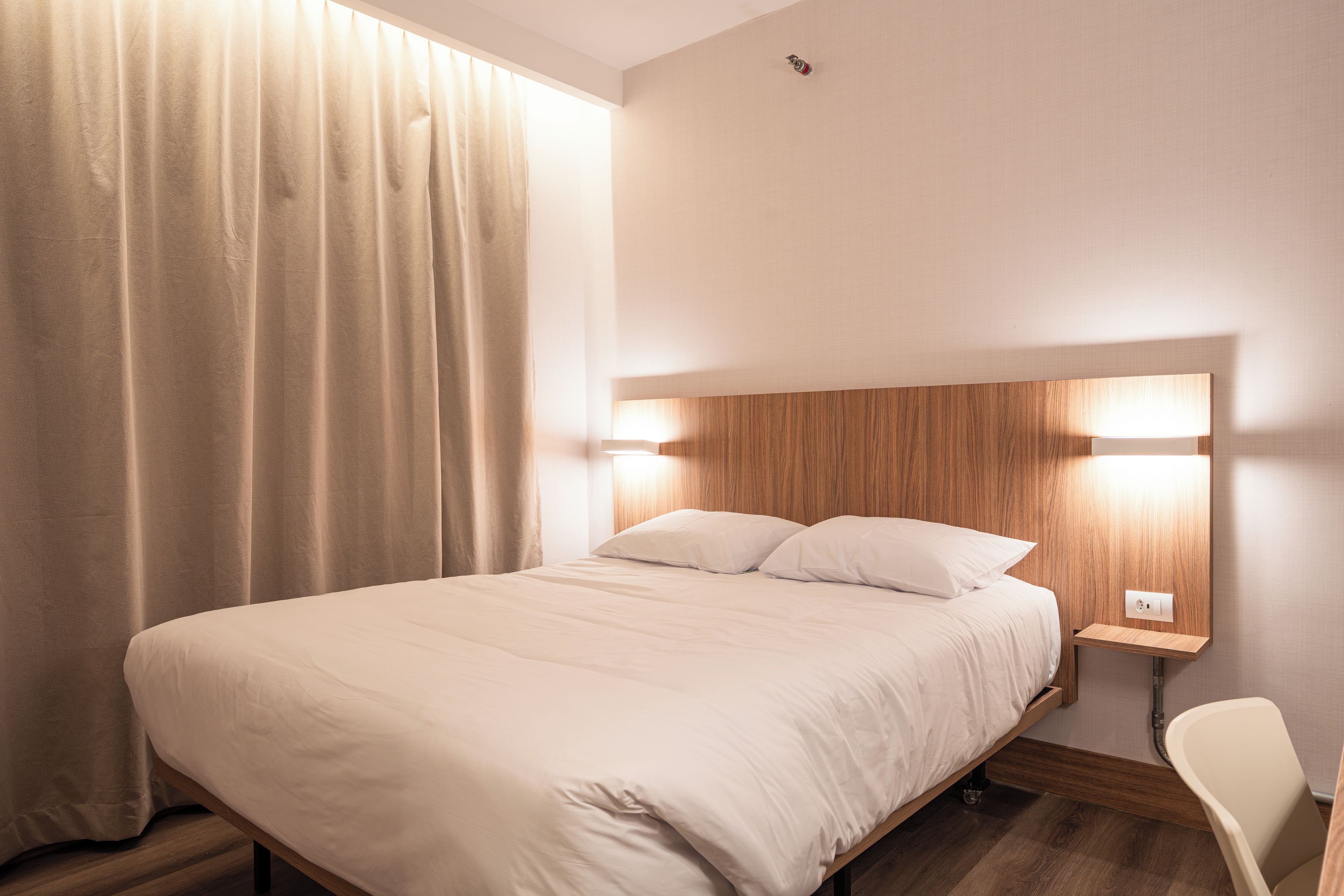 Hotellihuone, jossa on sänky, yöpöytä, lamppu, tuoli ja ikkuna, jossa on verhot.