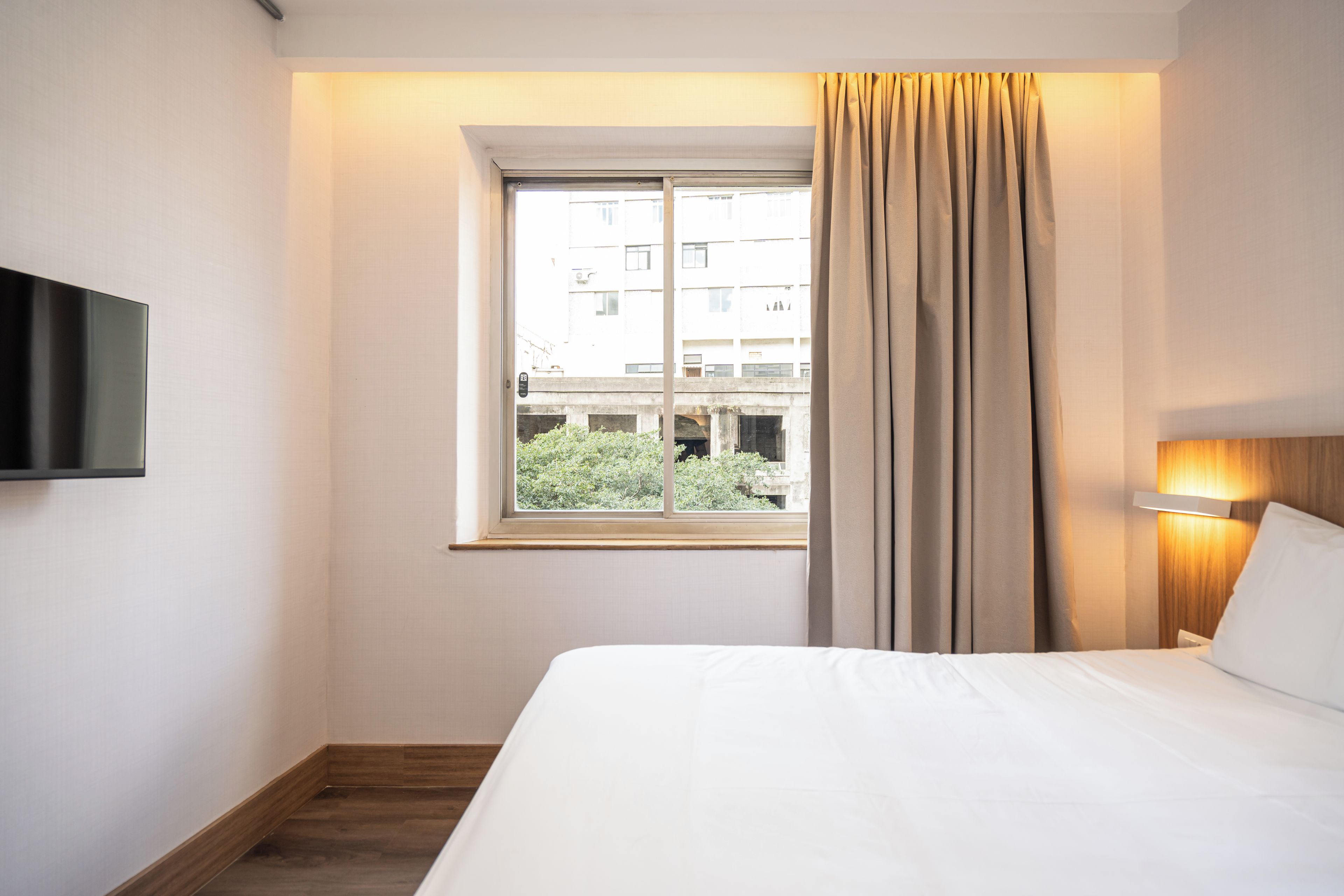 Habitación de hotel con una cama, un televisor en la pared, una ventana con cortinas y vistas a edificios.