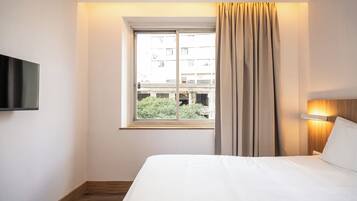 A hotel room with a bed, a television mounted on the wall, a window with curtains, and a view of buildings outside.