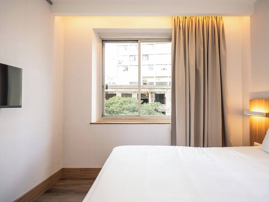 A hotel room with a bed, a television mounted on the wall, a window with curtains, and a view of buildings outside.