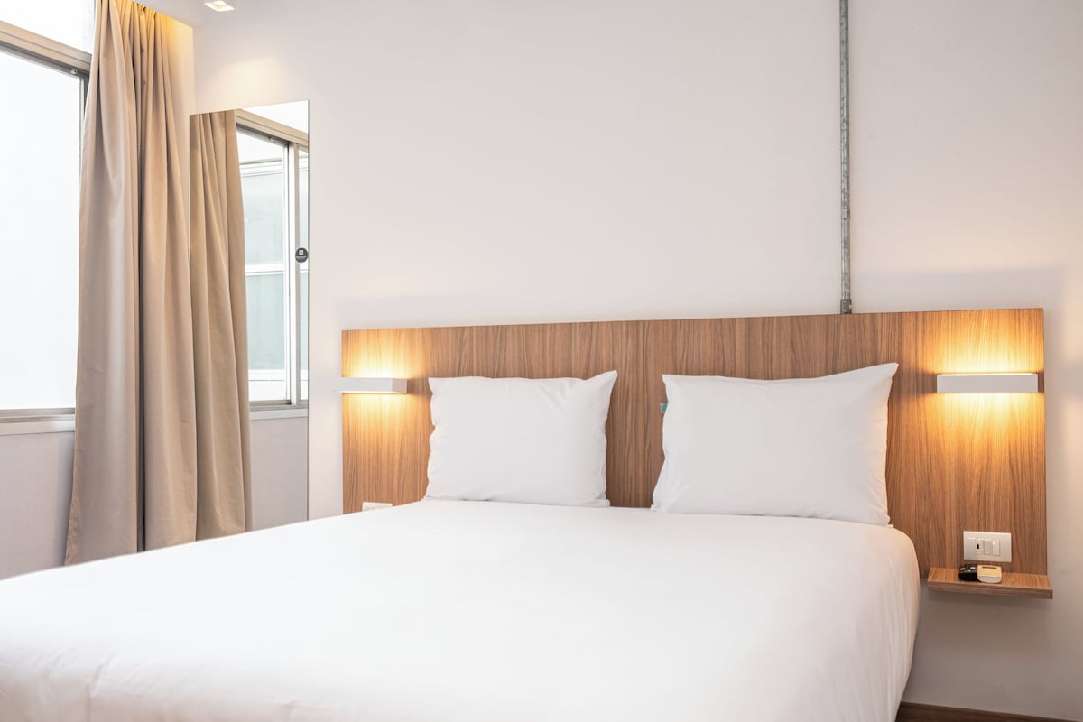 Una habitación de hotel con una cama, dos lámparas de noche, una mesita de noche y un ventanal con cortinas.