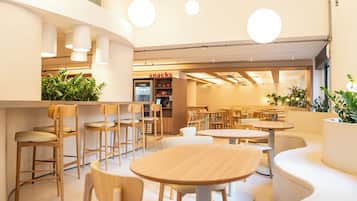 Un interno di un caffè moderno con tavoli rotondi e sedie di legno, un bancone per snack in vista e piante in vaso.