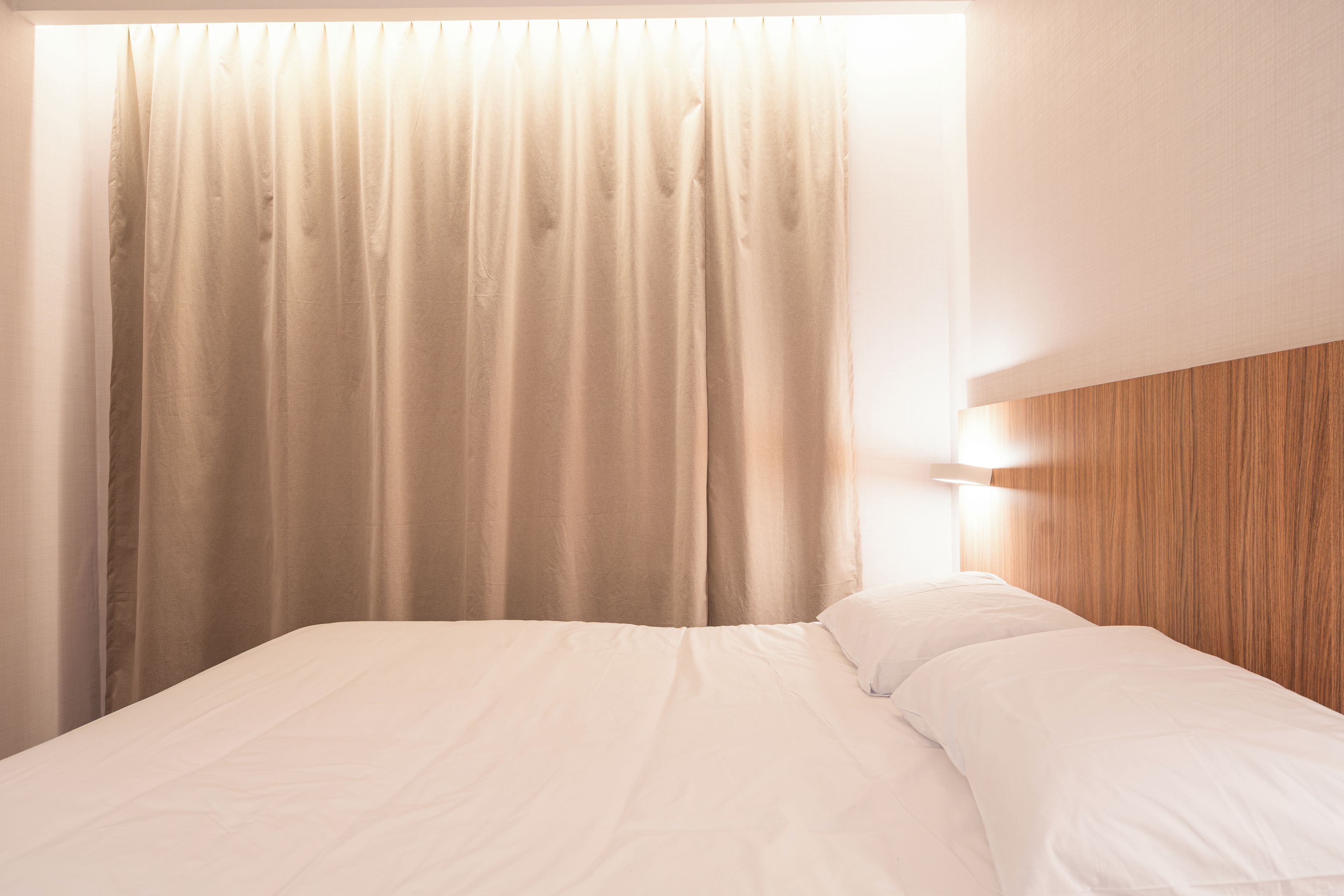 A bed with white bedding, a wooden headboard, and a window with beige curtains.