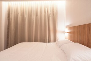 Una cama con ropa de cama blanca, cabecera de madera y ventana con cortinas color beige.