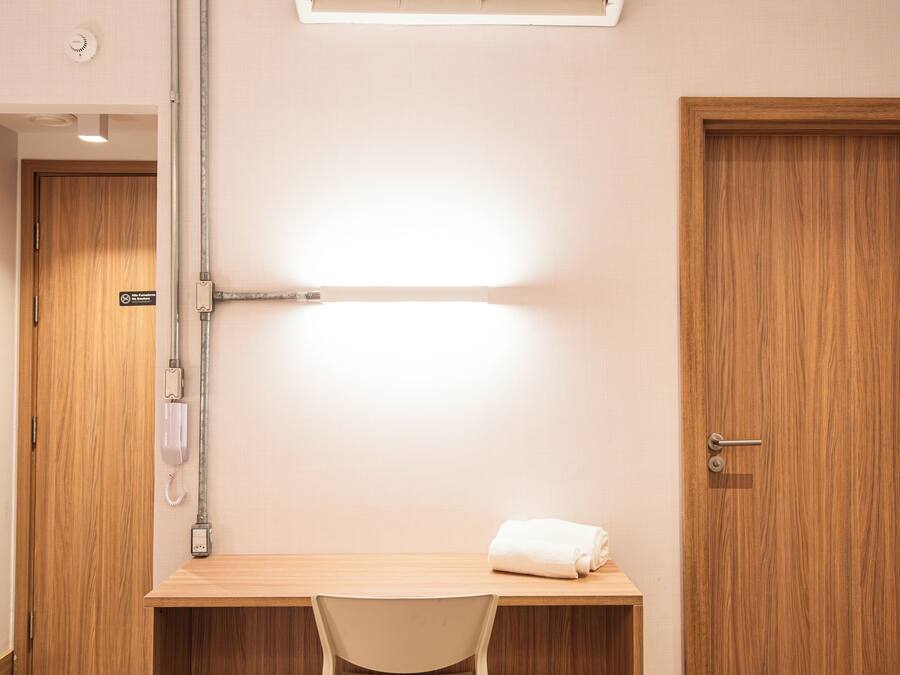A hotel room with a bed, a desk with a chair, an air conditioning unit, a telephone, and a door.
