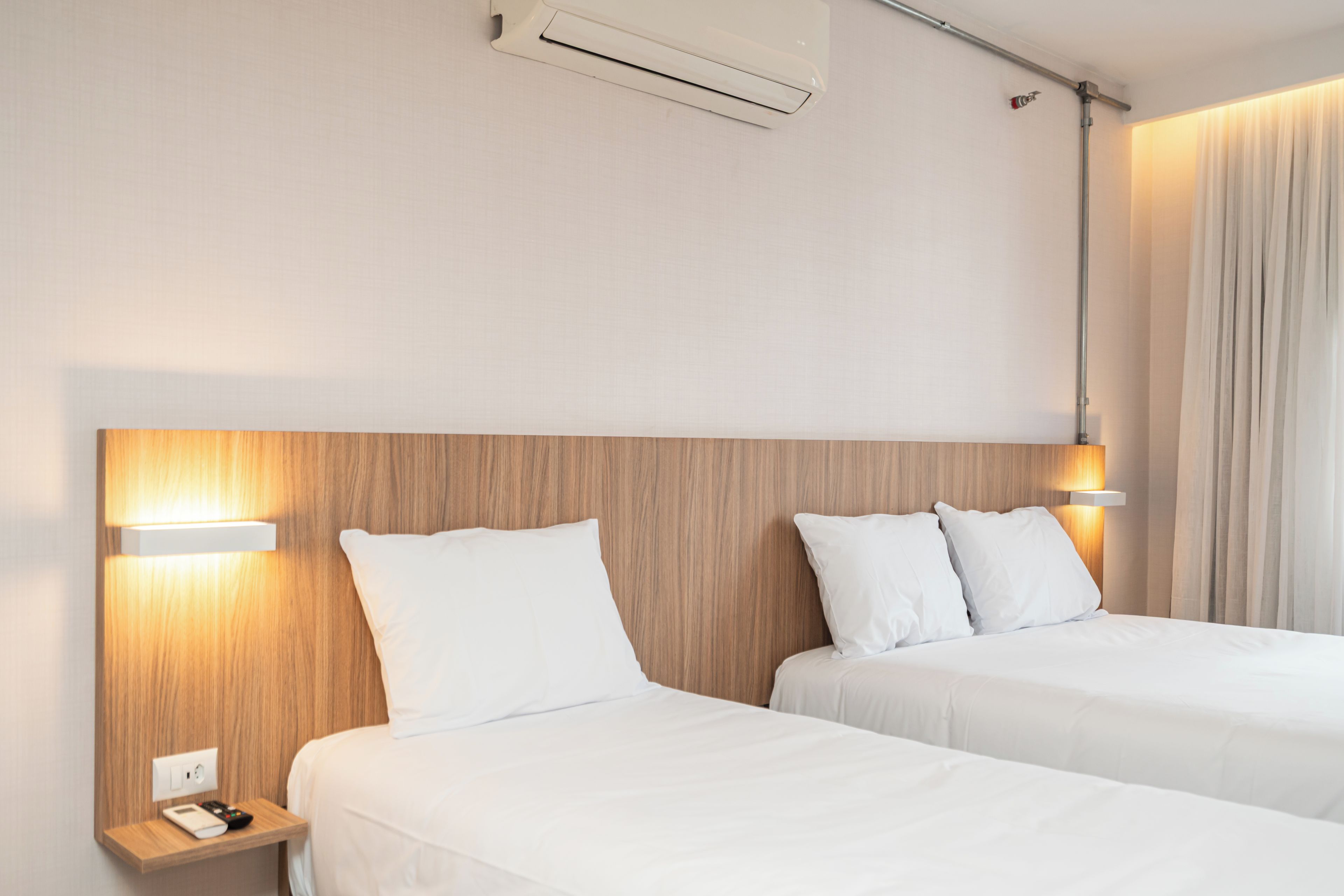 Quarto de hotel com duas camas, uma mesinha de cabeceira com telefone e controle remoto, e um aparelho de ar condicionado na parede.
