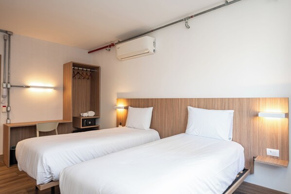 A hotel room with two beds, a wooden wardrobe, a desk with a chair, and a wall-mounted air conditioning unit.
