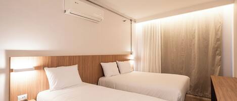 A hotel room with two single beds, a wooden headboard, a bedside table with a lamp, and a window with curtains.