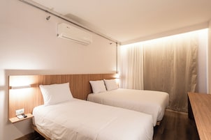 Hotellihuone, jossa on kaksi erillistä sänkyä, puinen pääty, yöpöytä lampun kanssa ja ikkuna, jossa on verhot.