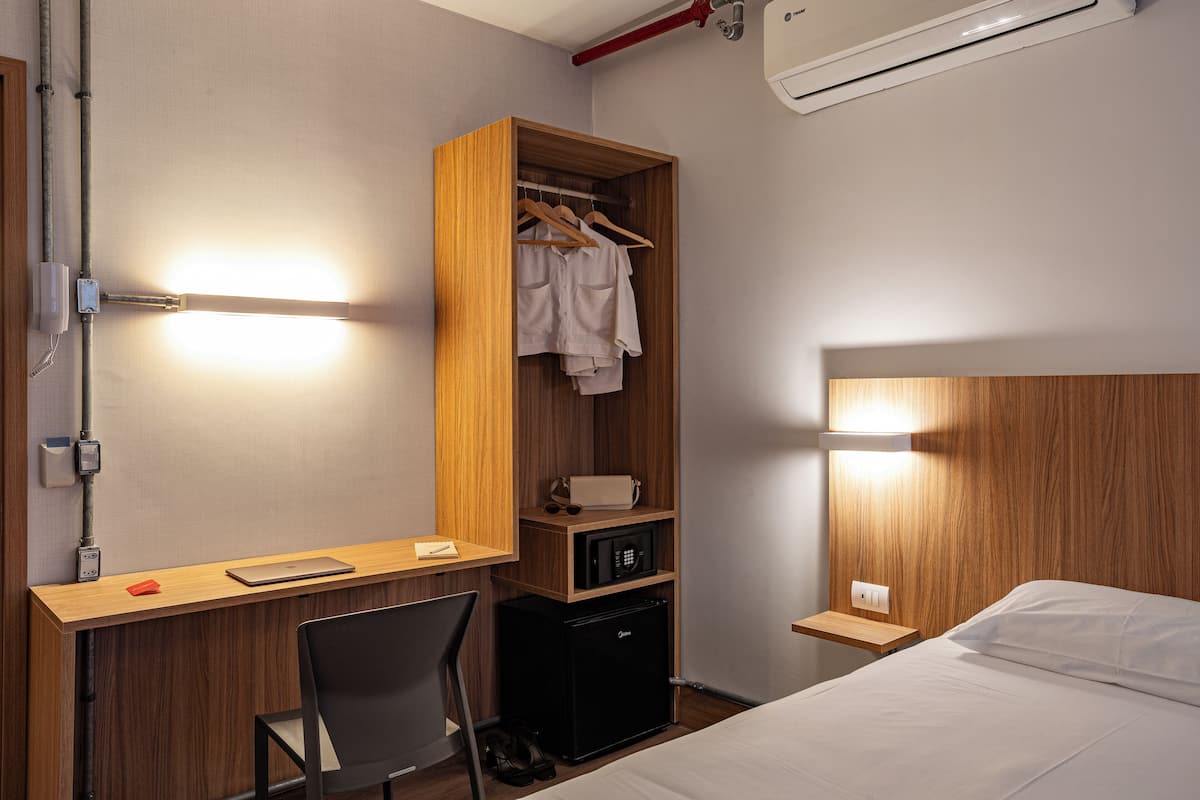 A hotel room with a wooden desk, a bed, a wardrobe, and a small refrigerator.