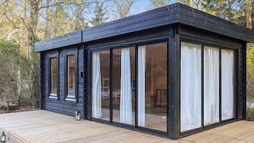 A modern cabin with large sliding glass doors, a deck, and a view of the surrounding forest.