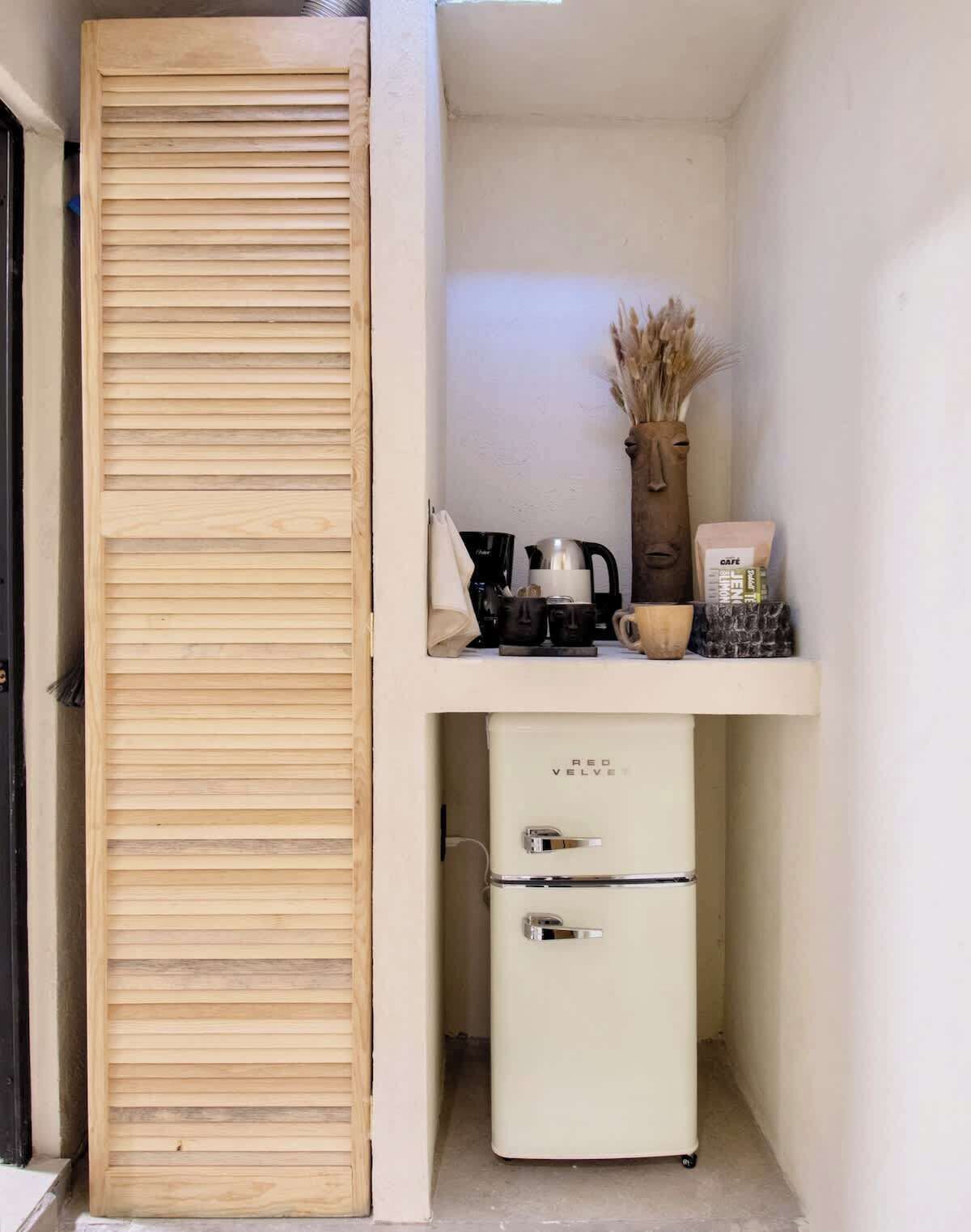 A compact kitchenette with a wooden sliding door, a vintage red velvet refrigerator, a coffee maker, and a vase with dried plants.