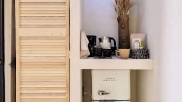 A compact kitchenette with a wooden sliding door, a vintage red velvet refrigerator, a coffee maker, and a vase with dried plants.