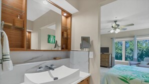Un baño moderno con un espejo grande, un lavamanos blanco y un mueble de tocador de madera. Hay un ventilador de techo y, en la habitación contigua, una cama con una colcha de estampado de hojas verdes.