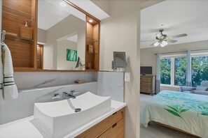 Ein modernes Badezimmer mit einem großen Spiegel, einem weißen Waschbecken und einer Holzanrichte. Im angrenzenden Raum befindet sich ein Deckenventilator und ein Bett mit Bettwäsche in Grün- und Blattmuster.