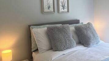 Een bed met zachte kussens, een bedlampje en twee ingelijste botanische afbeeldingen aan de muur.