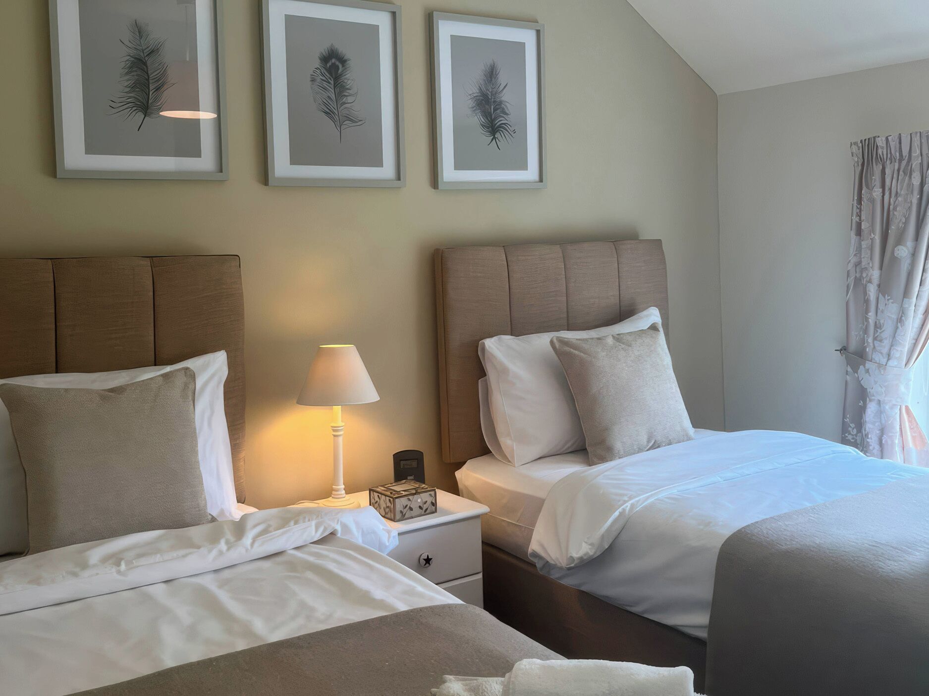 Habitación de hotel con dos camas, una mesita de noche con lámpara y tres cuadros con motivos de plumas en la pared.