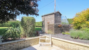 Una cabaña de madera con un estante de metal y una olla colgante, rodeada por un jardín con un muro de ladrillo y una reja metálica.