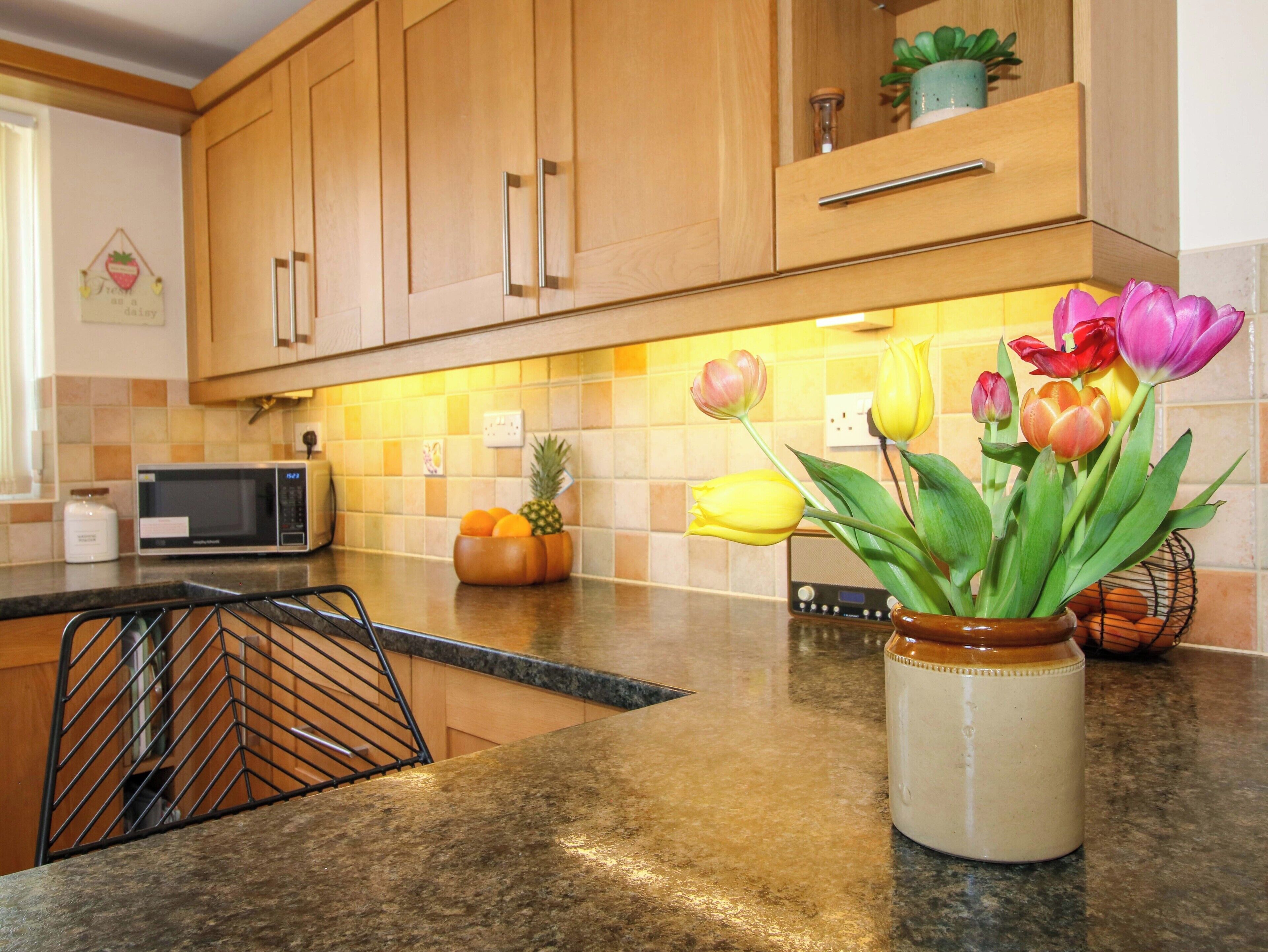 Una cocina con armarios de madera, un microondas, un frutero y un jarrón con flores.