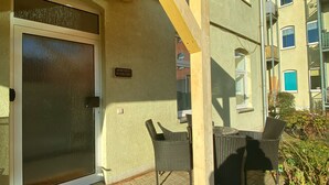 A covered patio with a sliding glass door, outdoor seating, and a brick pathway leading to a building entrance. - Apartment at the ford (Erfurt)