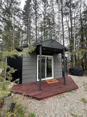 Una piccola dependance con una porta vetrata, circondata da una terrazza e ghiaia, immersa in una foresta.