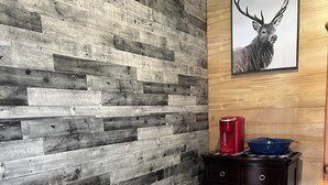 Una habitación con pared de madera, chimenea, una silla de madera, cómoda y un cuadro enmarcado de un ciervo.