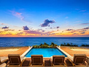 Una piscina con sillas de descanso frente al océano al atardecer.