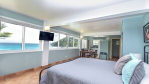 A bedroom with a large bed, a TV mounted on the wall, a dining area, and a view of the ocean. - The Breeze (ST Thomas)