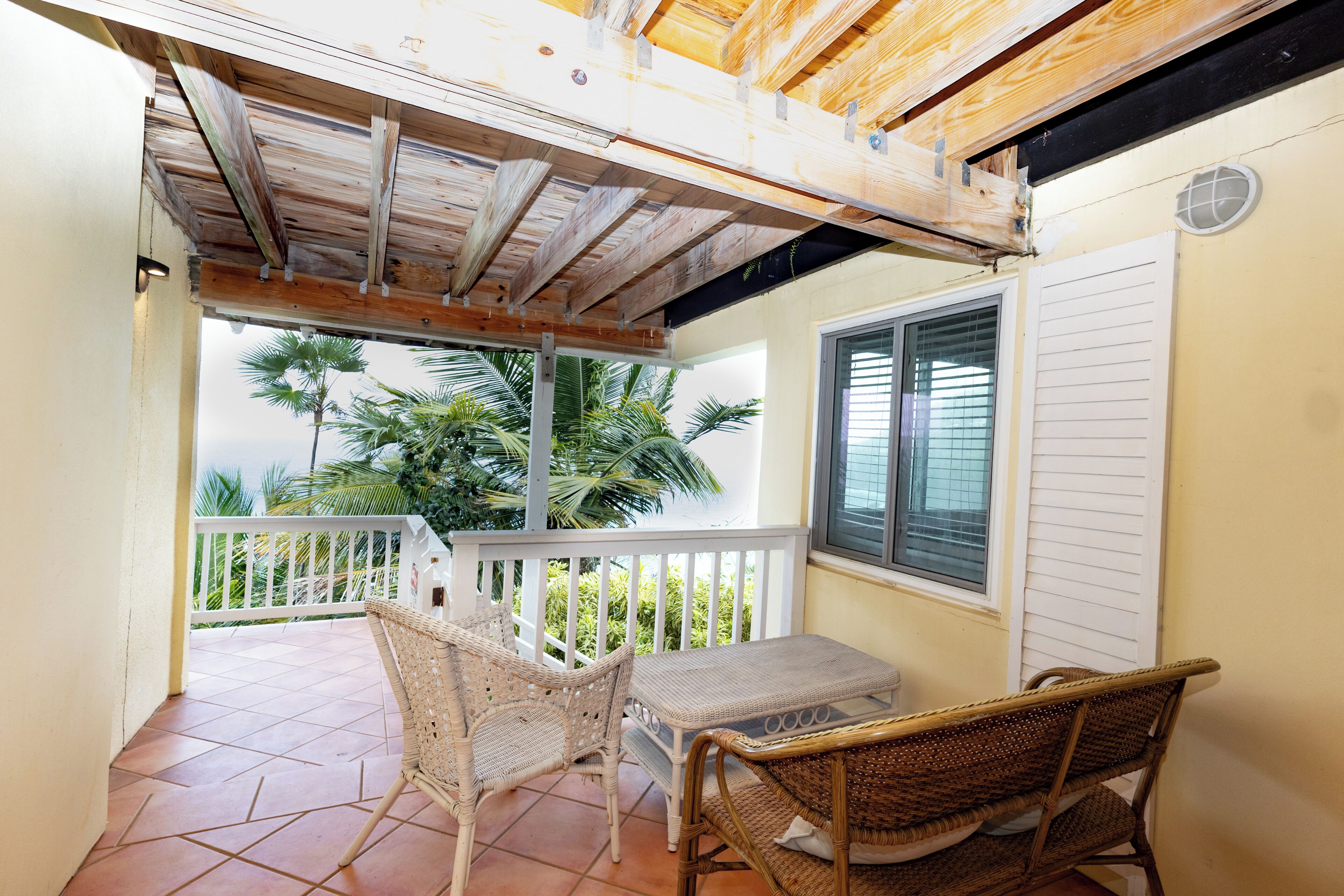 A covered outdoor seating area with wicker furniture, a tiled floor, and a view of palm trees.