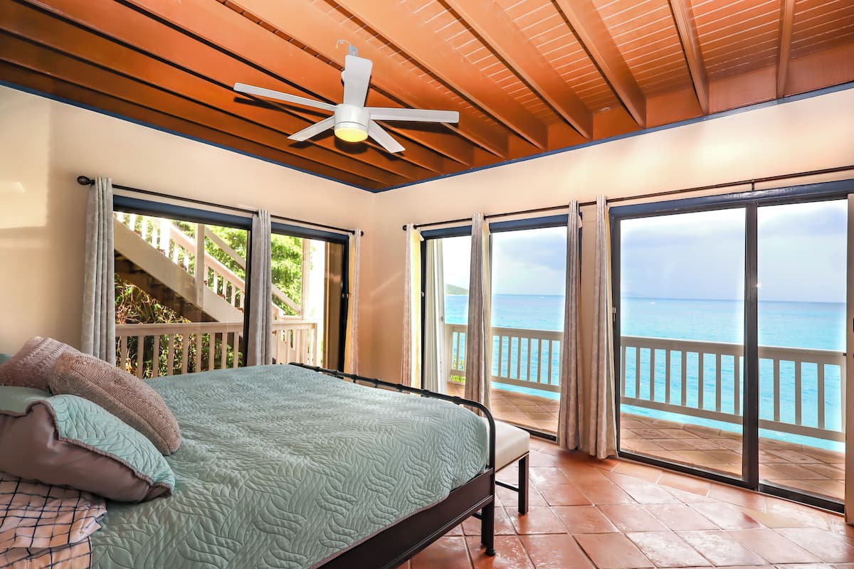 A bedroom with a bed, a ceiling fan, and a view of the ocean through large sliding doors.