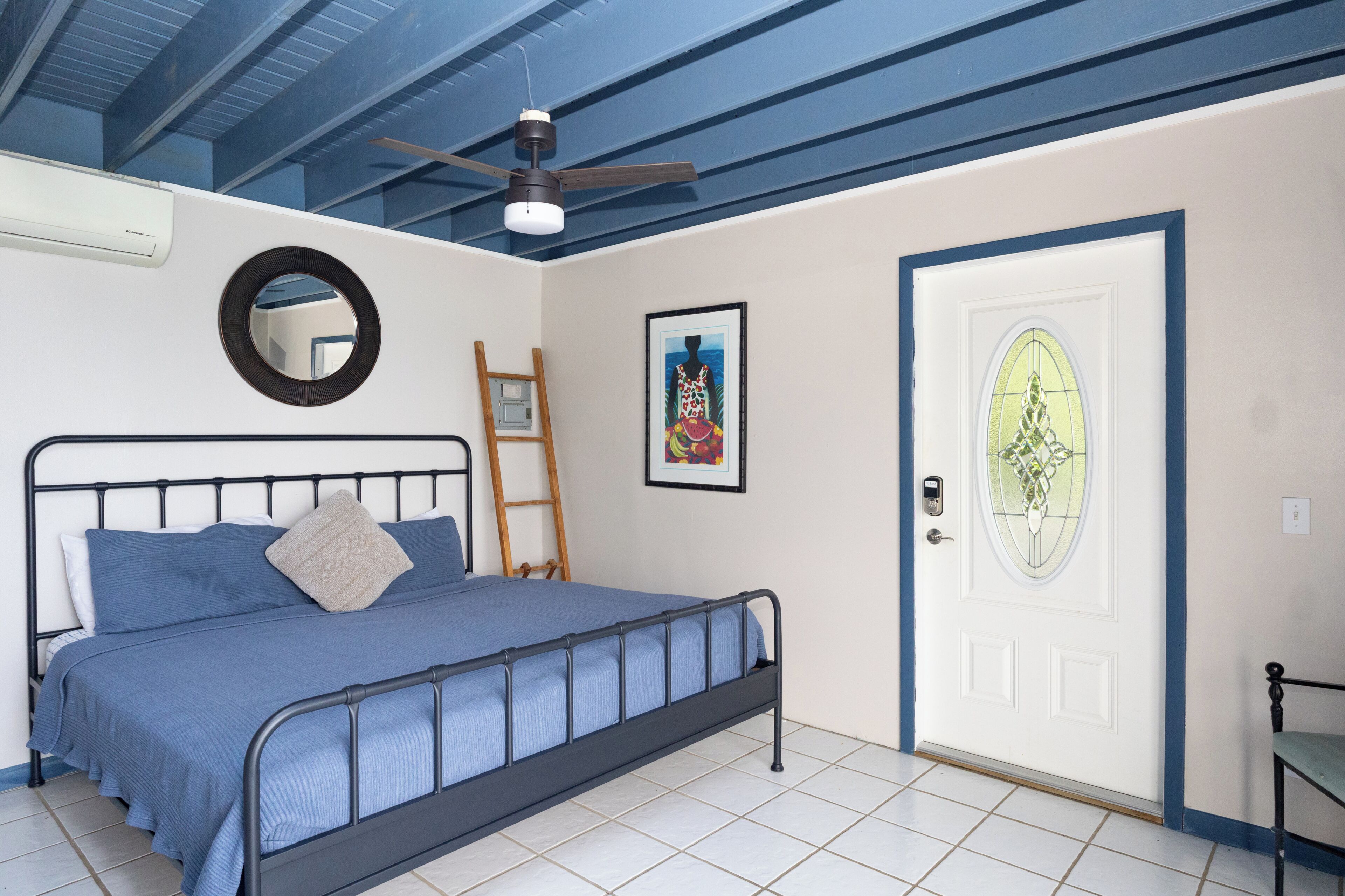A bedroom with a bed, a ceiling fan, a door with a glass panel, a framed picture on the wall, and a ladder.