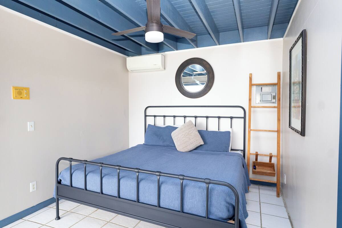 A bedroom with a bed, a ceiling fan, an air conditioning unit, a small wooden ladder, and a framed picture on the wall.