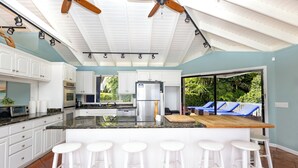 A modern kitchen with white cabinets, a central island with bar stools, a ceiling fan, and a view of a backyard with a pool. - The Breeze (ST Thomas)