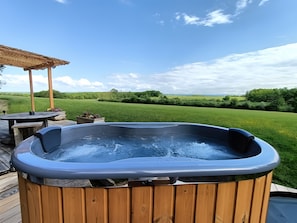 A hot tub on a wooden deck with a view of a grassy field and a clear sky.