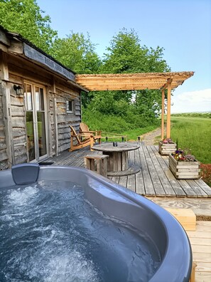 Una cabaña rústica de madera con jacuzzi, terraza de madera, mobiliario exterior y una pérgola.